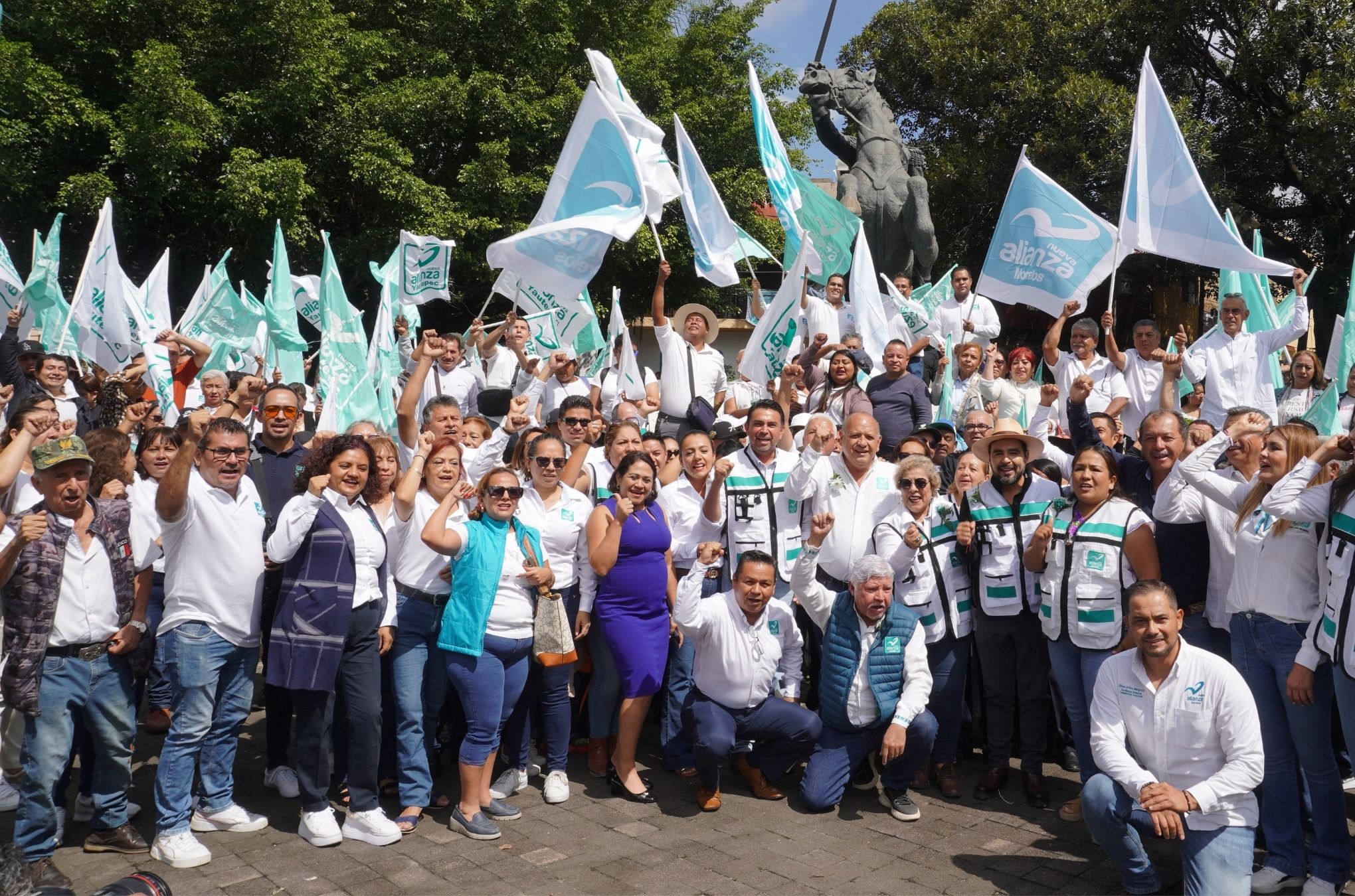 Militantes de Nueva Alianza Morelos con banderas del partido reunidos en una plaza pública frente al monumento a Emiliano Zapata.